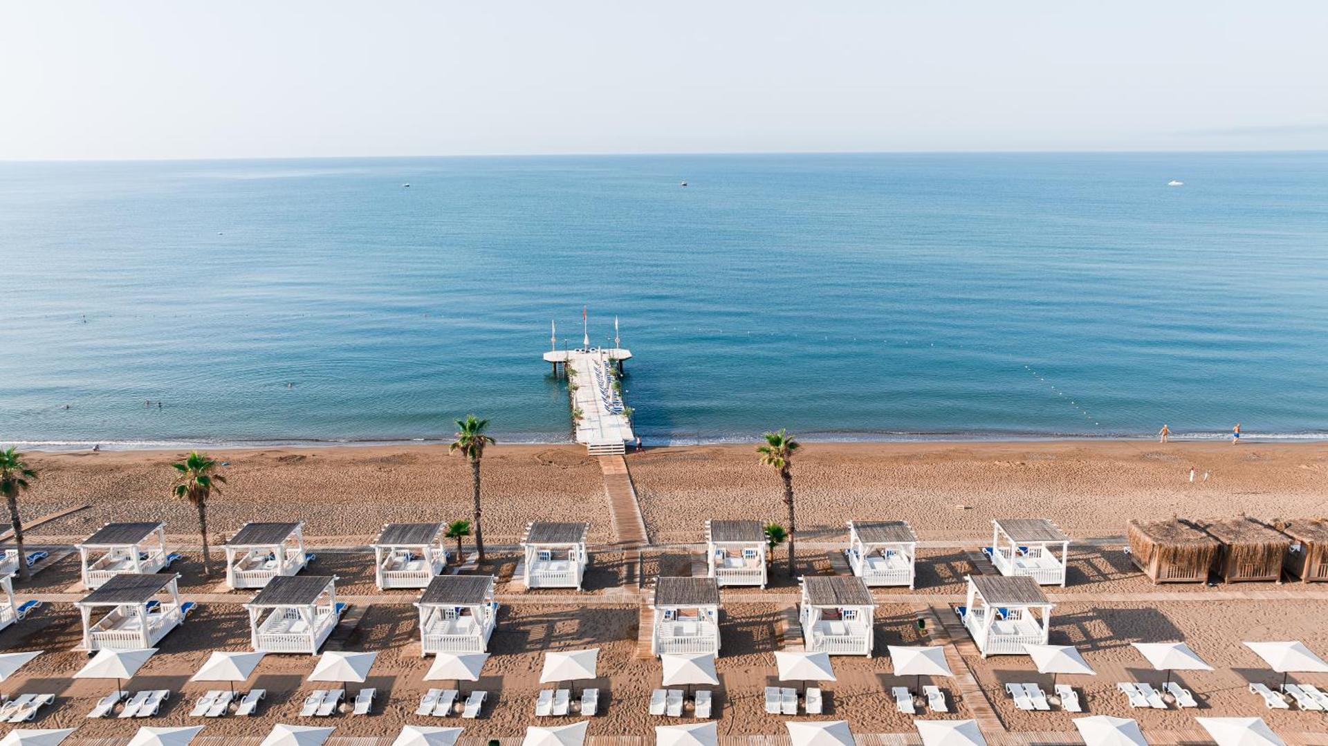 Adora Hotel & Resort privātā smilšu pludmale Belekā — koka piestātne, balti pludmales bungalo un sauļošanās krēsli pie zilas jūras