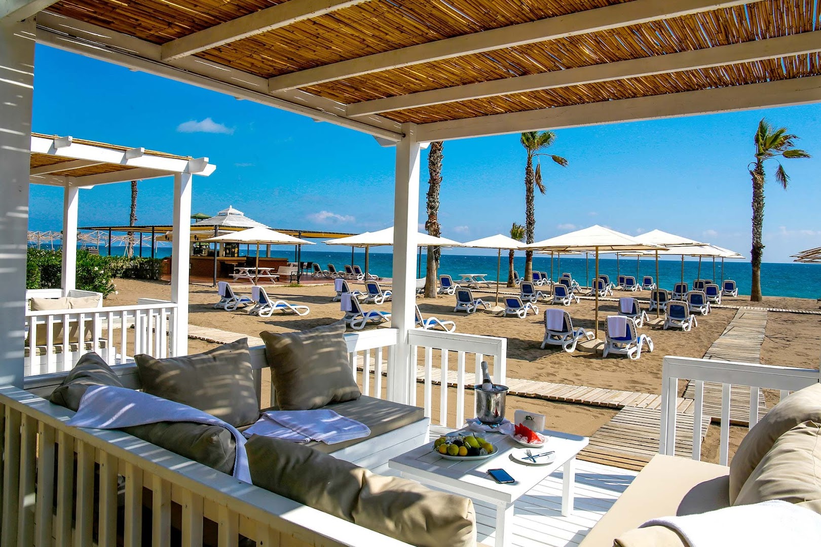 Adora Hotel & Resort atpūtas zona pludmales bungalo iekšpusē — balti dīvāni, galdiņš ar augļiem un skats uz jūru un sauļošanās krēsliem