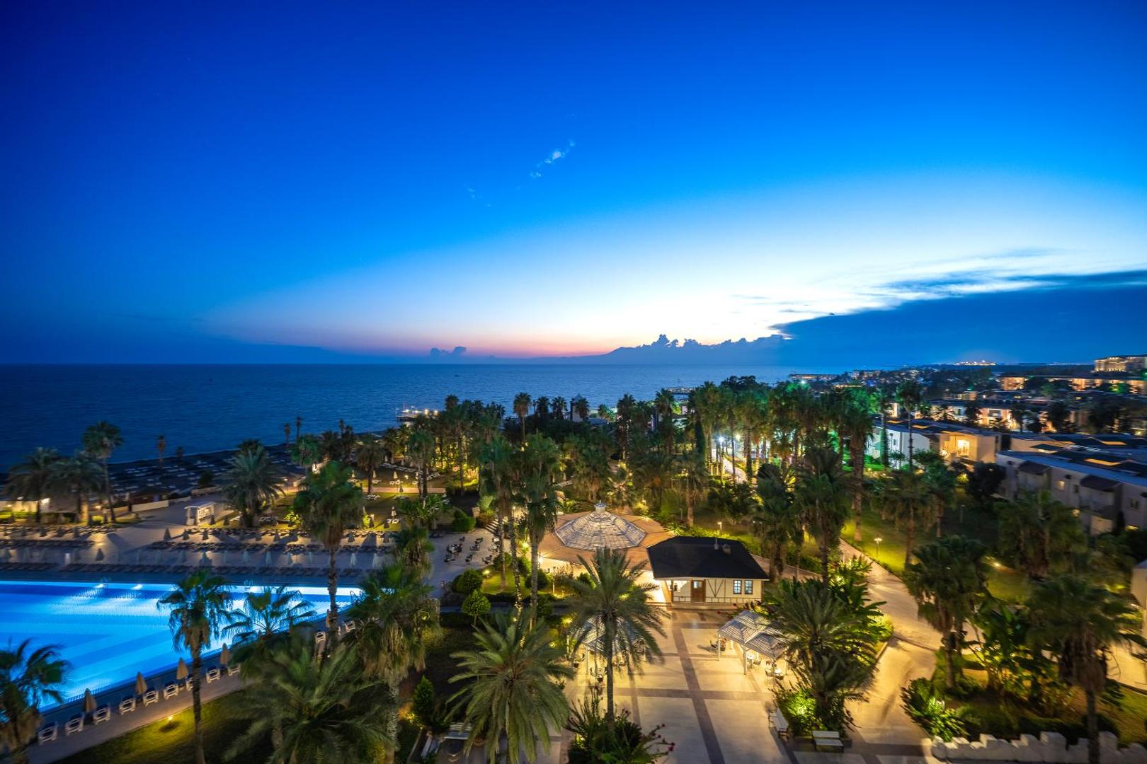 Adora Hotel & Resort vakara apgaismojums dārzā un pie baseina — palmu ieskauta teritorija un skats uz jūru krēslas stundā