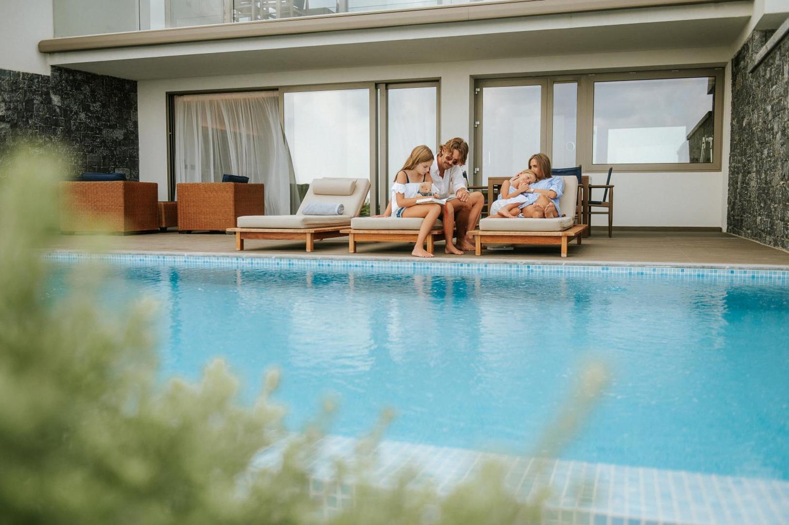 Ģimene atpūšas pie privātā baseina Nana Princess Suites Villas & Spa kūrortā Krētā — sauļošanās krēsli un tieša piekļuve ūdenim