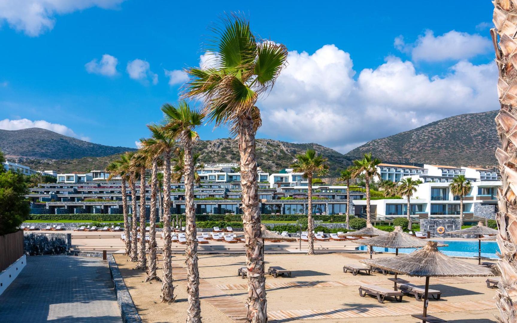 Nana Princess Suites Villas & Spa pludmales zona ar palmām, saulessargiem un kalnu fonu Krētas piekrastē