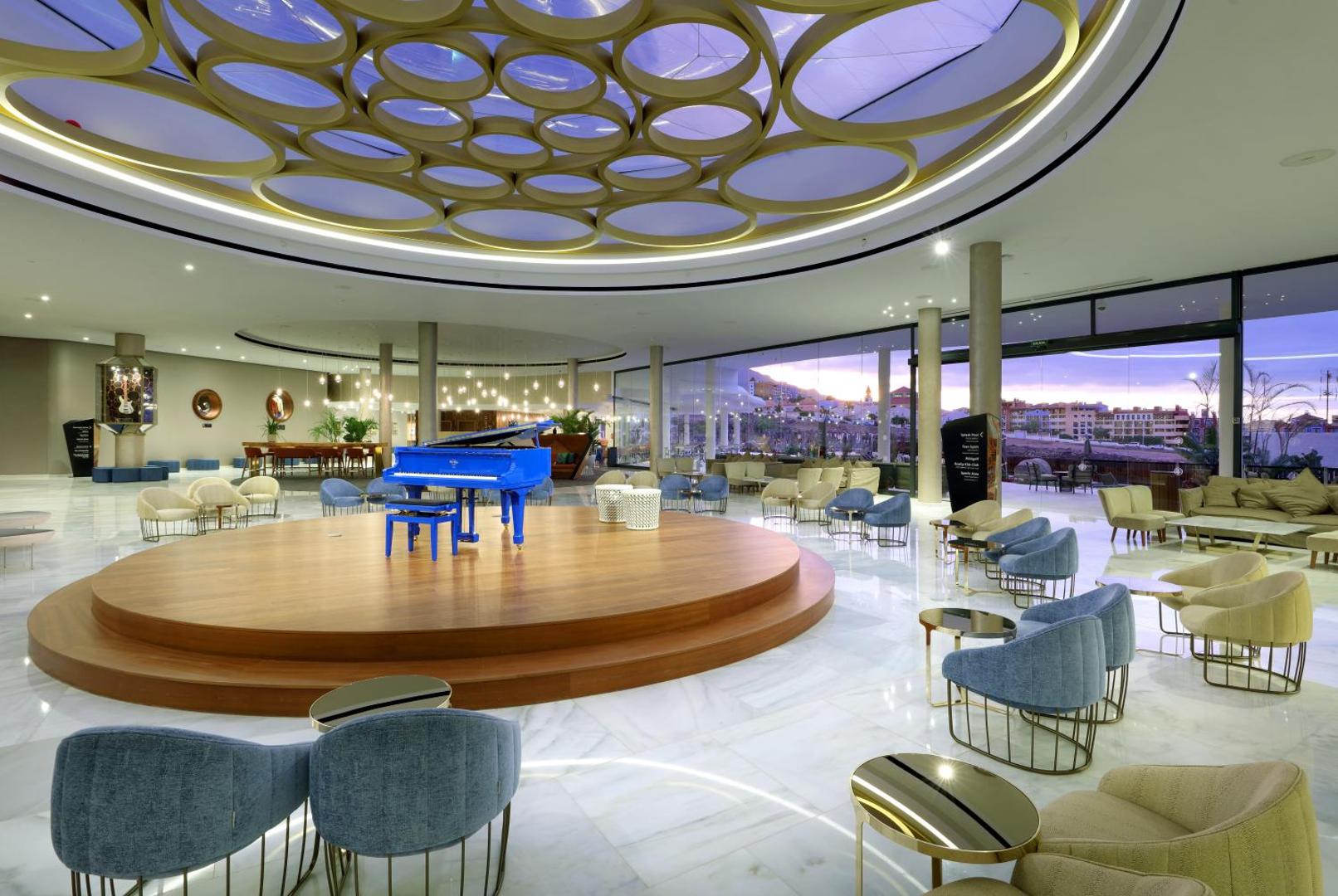 Hard Rock Hotel Tenerife plašā vestibila atpūtas zona ar zilu flīģeli centrā, modernu griestu dizainu un skatu uz pilsētu saulrietā.