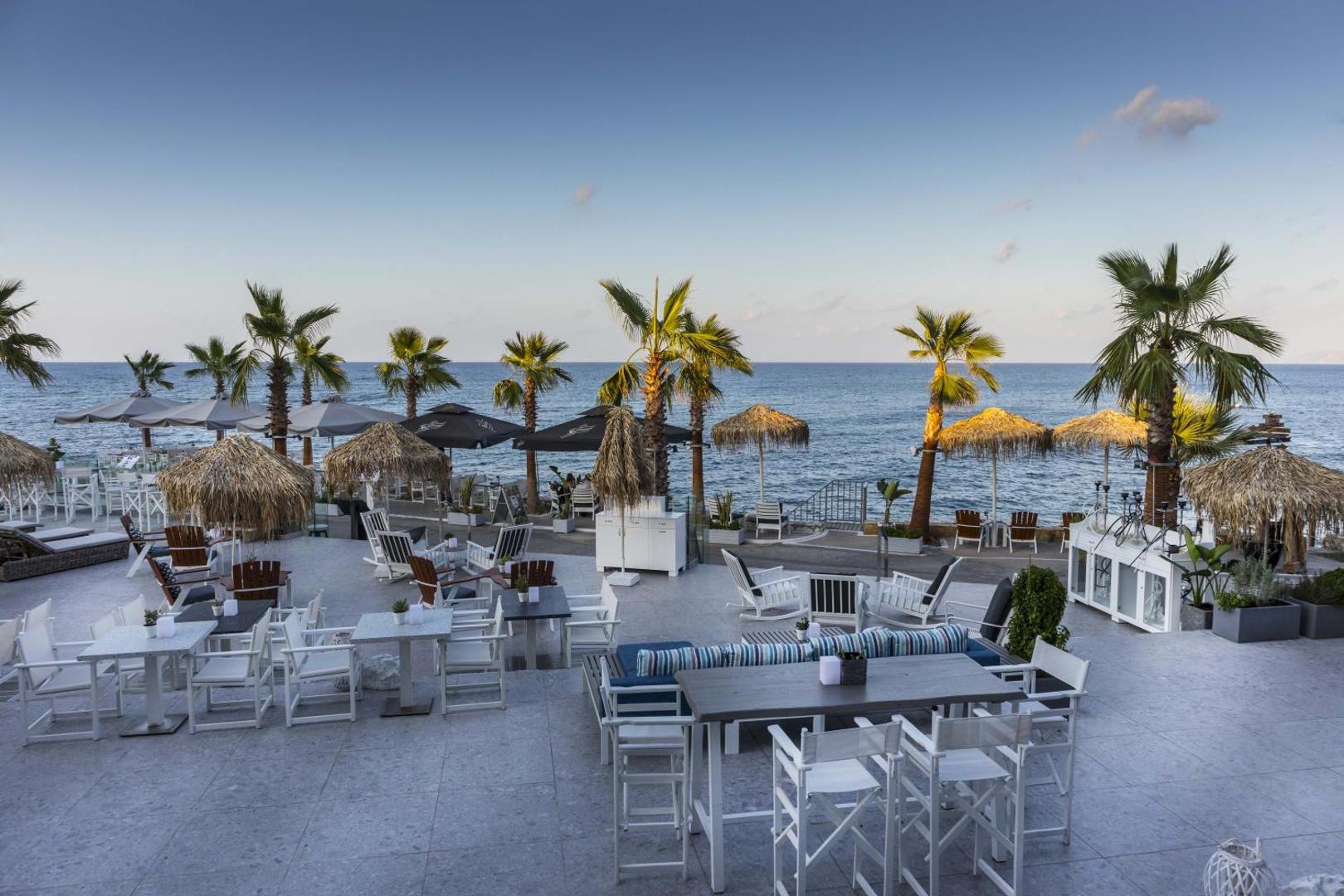 Glaros Royal Beach viesnīcas āra terase ar skatu uz jūru Hersonisā, Krētā — balti krēsli, palmas un saulessargi piekrastē