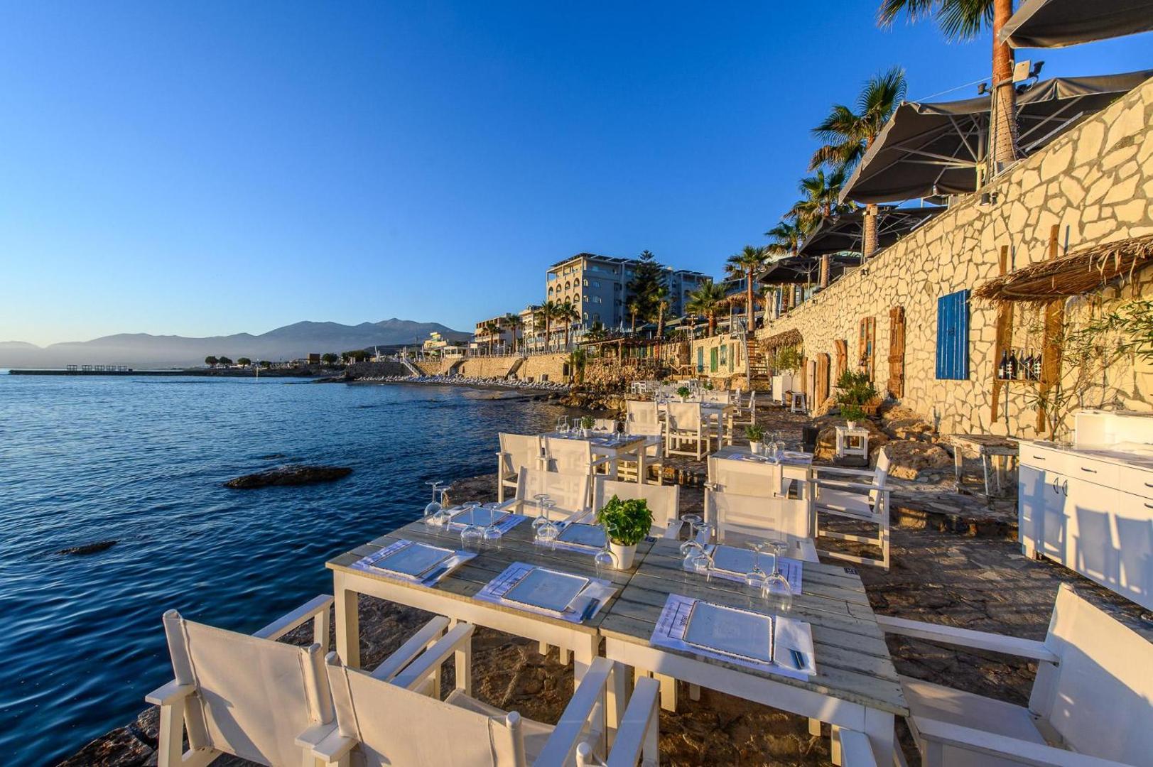 Glaros Royal Beach ēdienkartes restorāns uz akmens terases pie jūras Hersonisā — balti galdi un krēsli saulrieta gaismā