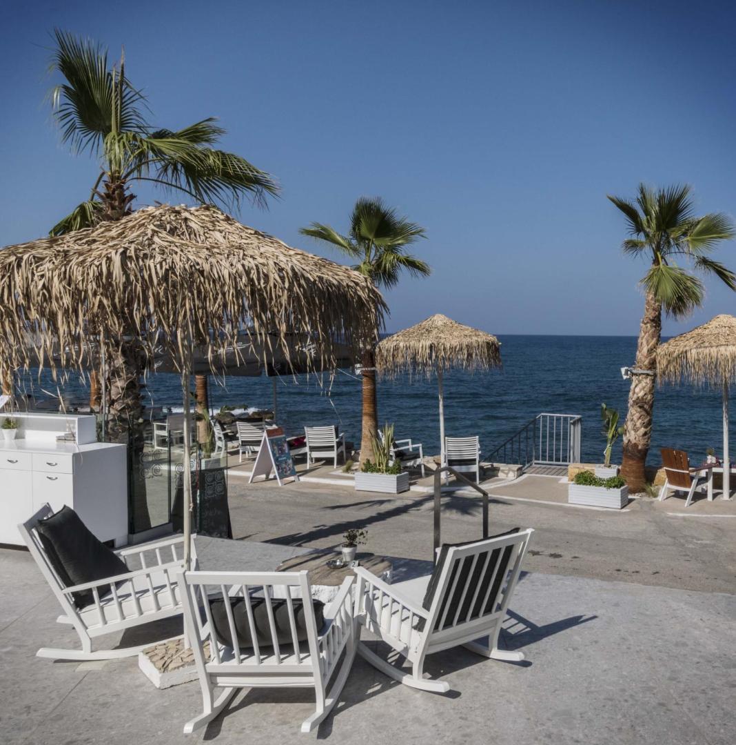 Glaros Royal Beach viesnīcas atpūtas zona Hersonisā, Krētā — balti šūpuļkrēsli zem salmu saulessargiem pie pašas jūras krasta līnijas