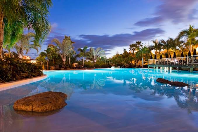 Bahia del Duque āra baseins vakara apgaismojumā ar izgaismotām palmām un tiltiņu Tenerifes kūrortā