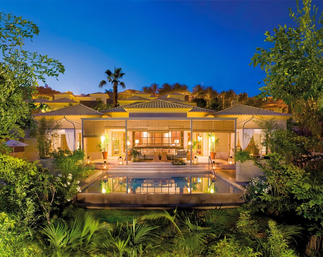 Bahia del Duque grezna bungalo tipa villa ar privātu baseinu, terasi un tropisku dārzu vakara apgaismojumā.