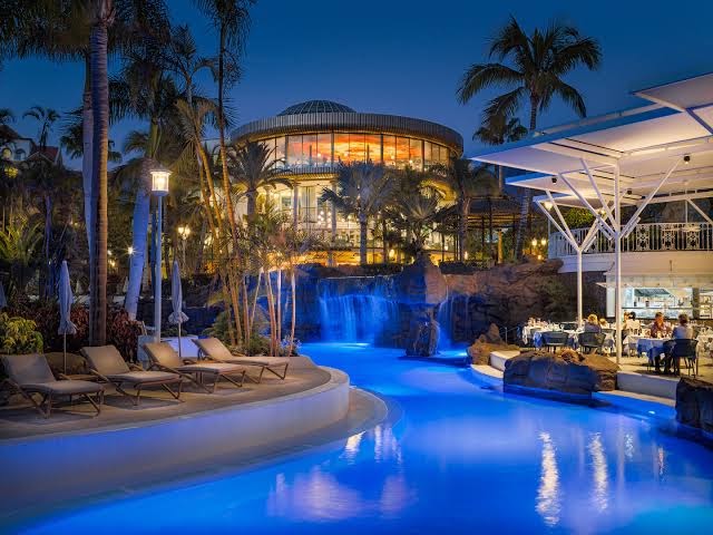 Jardines de Nivaria vakara apgaismojums pie baseina ar ūdenskritumu, restorāna terasi un izgaismotu kupola ēku Tenerifē.