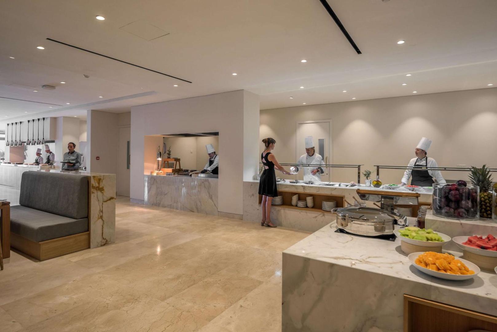 Cap St Georges Hotel & Resort bufetes tipa restorāns ar marmora apdari un pavāriem, kas apkalpo viesus pie atvērtās virtuves