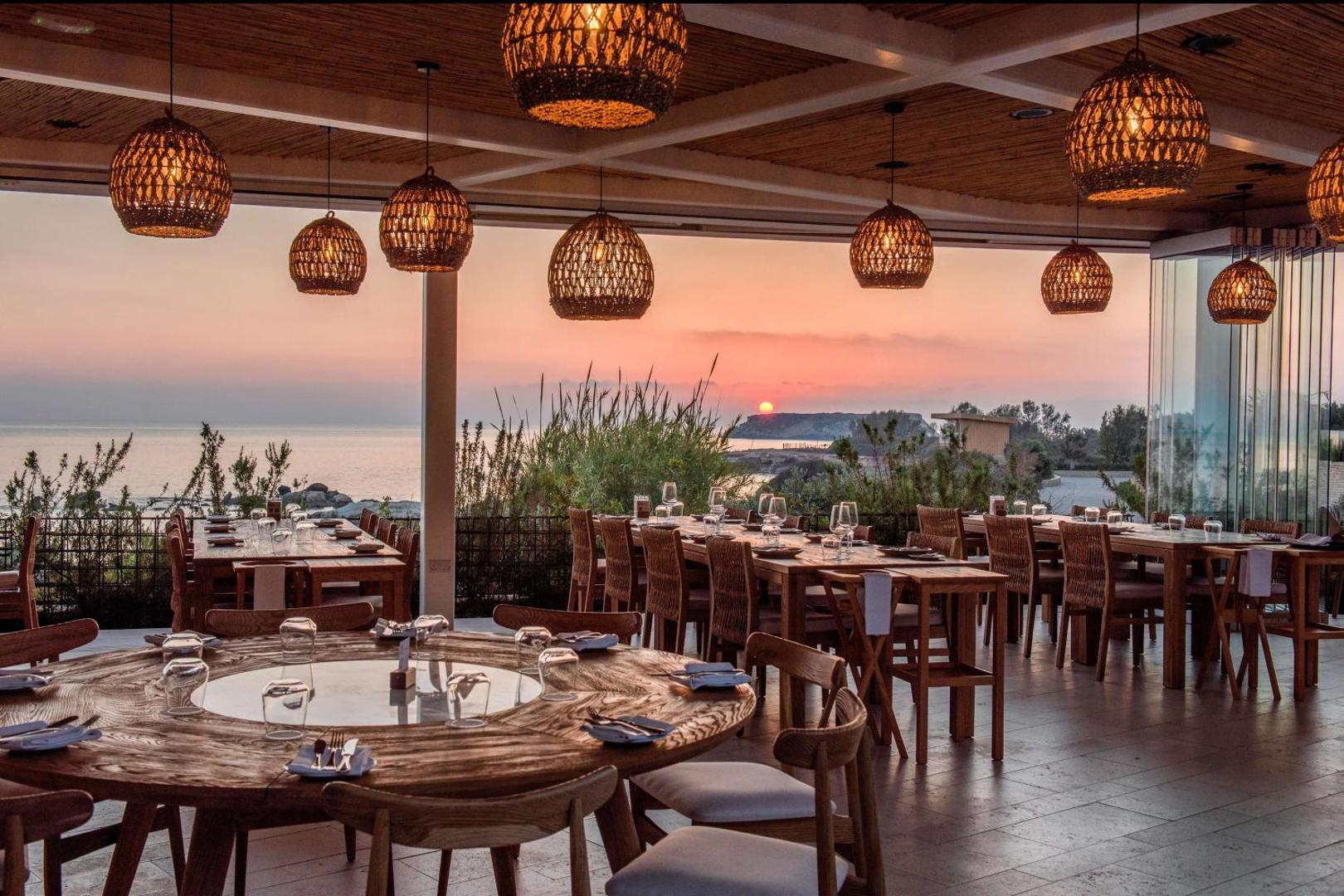 Cap St Georges Hotel & Resort restorāna terase ar koka mēbelēm un pītiem gaismekļiem saulrieta laikā Kipras piekrastē