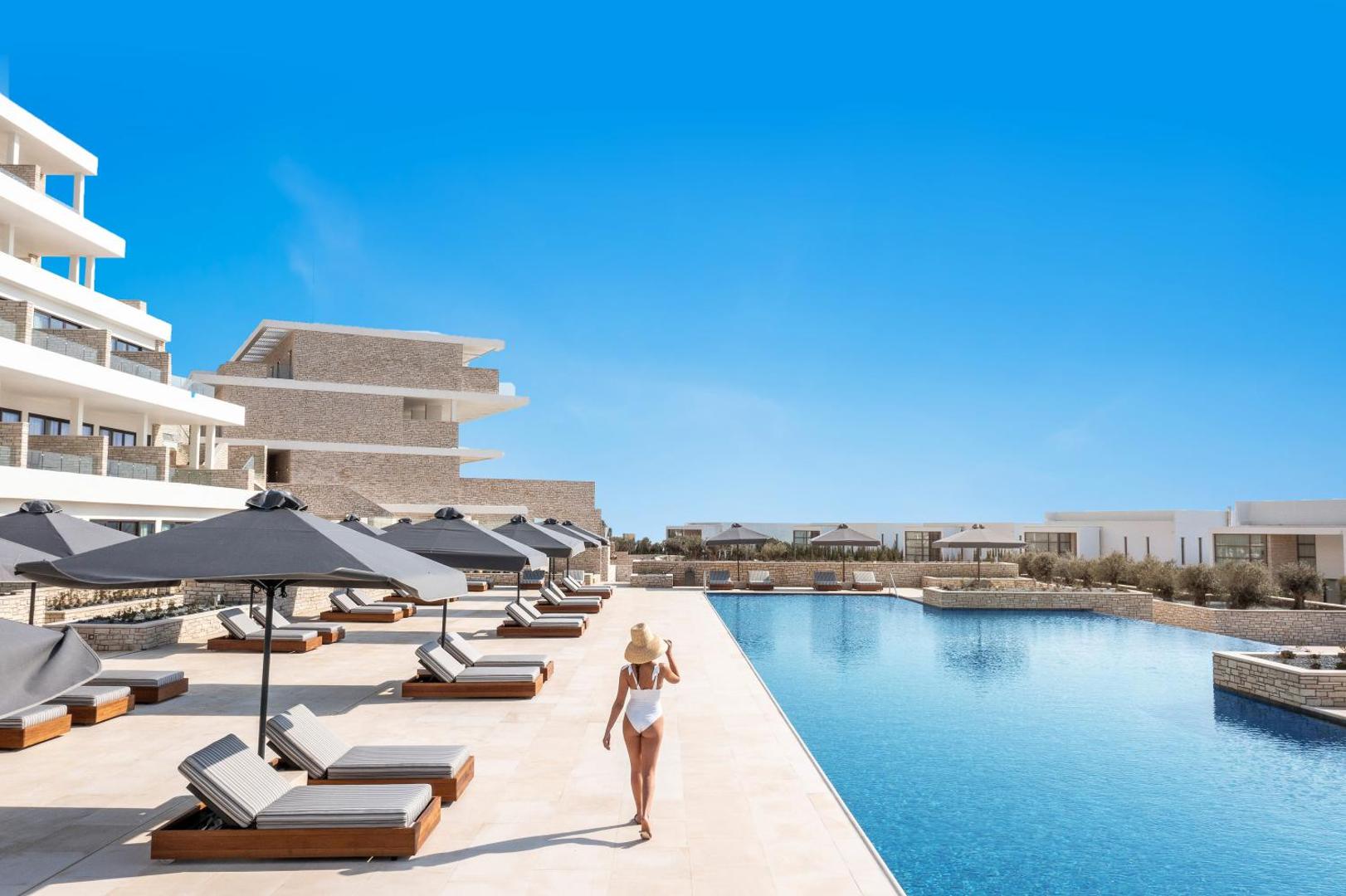 Cap St Georges Hotel & Resort lielais āra baseins ar sauļošanās krēsliem, saulessargiem un modernu arhitektūru Pafas reģionā