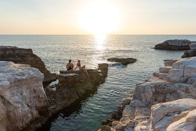Pāris vēro saulrietu uz baltajām klintīm pie Cap St Georges Hotel & Resort Kipras piekrastē — romantiska atmosfēra pie jūras