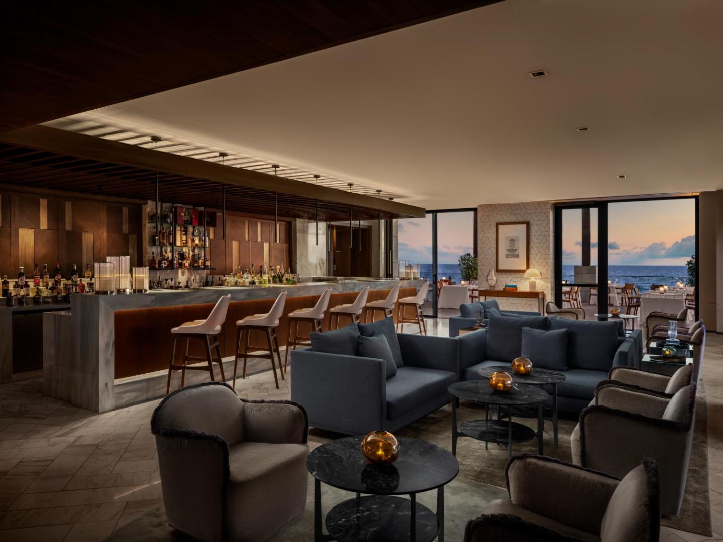 Amara Hotel lobby bārs ar zilajiem dīvāniem, marmora leti un panorāmas logiem uz jūru krēslas stundā