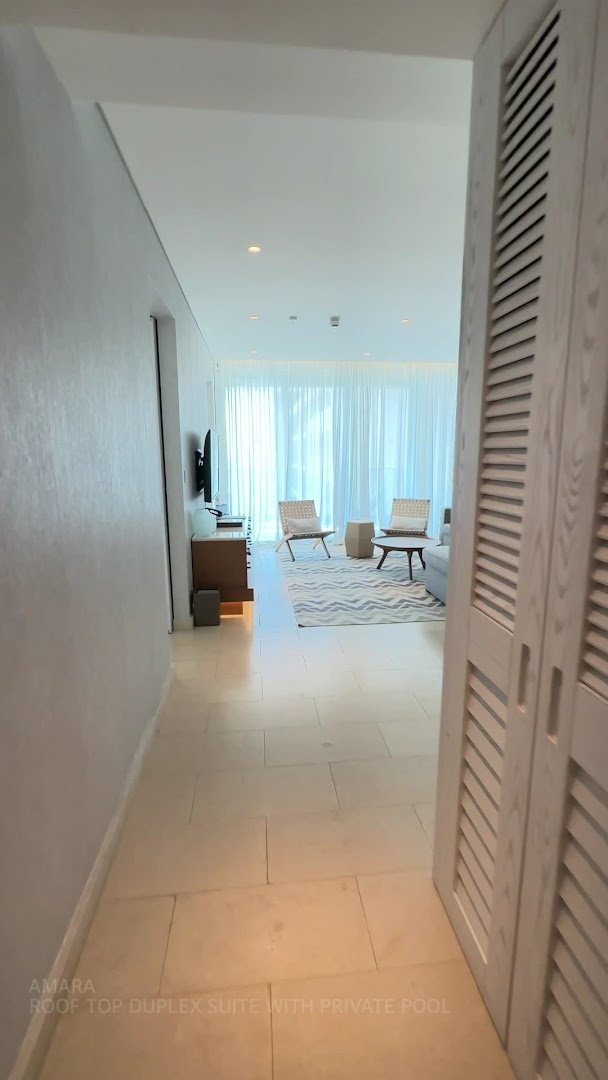 Amara Hotel Roof Top Duplex Suite dzīvojamā zona ar gaišām mēbelēm, paklāju un skatu uz terasi caur caurspīdīgiem aizkariem.