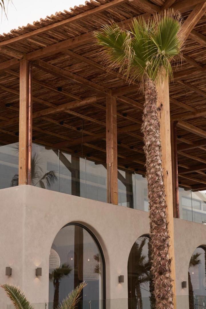 Stella Blue Seaside Resort viesnīcas fasādes detaļa ar koka konstrukcijām, arkveida logiem un augstām palmām saulainā dienā