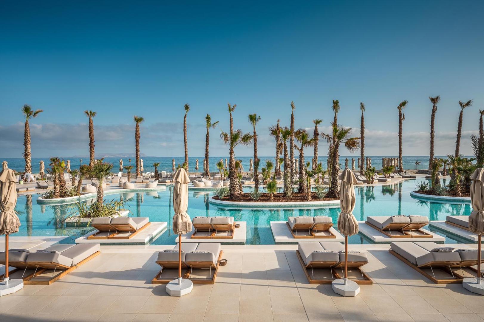 Stella Blue Seaside Resort plašais āra baseins ar sauļošanās krēsliem ūdenī, palmām un skatu uz jūru saulainā dienā.