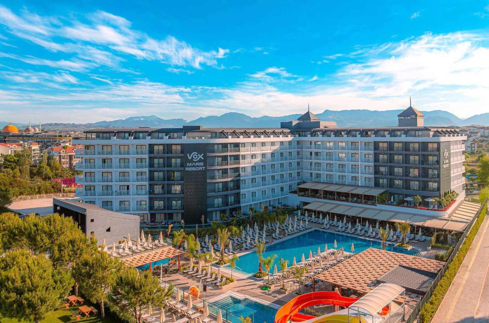 Panorāmas skats uz Vox Maris Resort viesnīcu, baseiniem un ūdens slidkalniņiem uz kalnu fona saulainā dienā Sidē