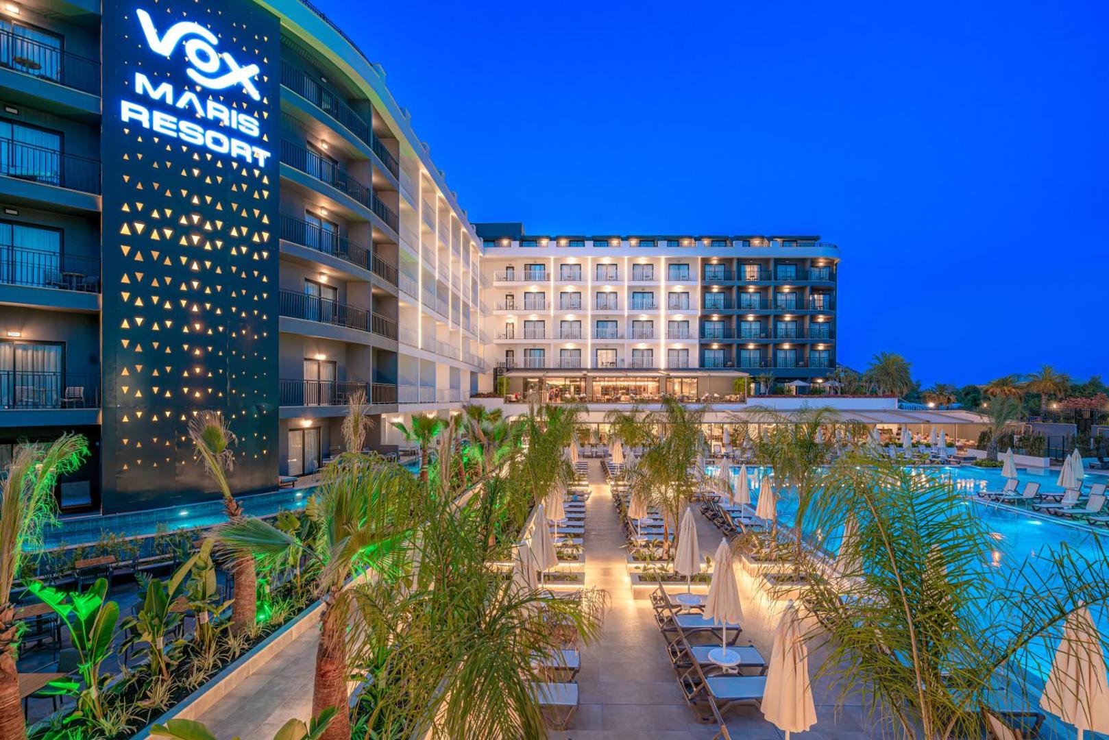 Vox Maris Resort fasāde un baseina zona zilajā stundā — izgaismotas palmas un terases Sides kūrortā