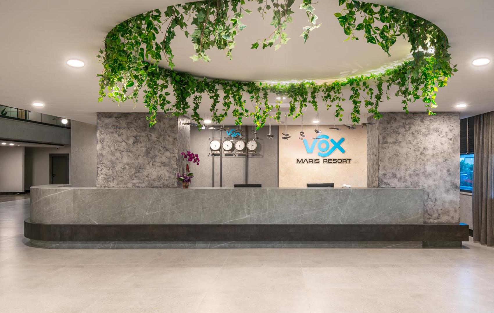 Vox Maris Resort viesnīcas recepcijas zona ar modernu leti, dekoratīviem augiem un pasaules pulksteņiem pie sienas