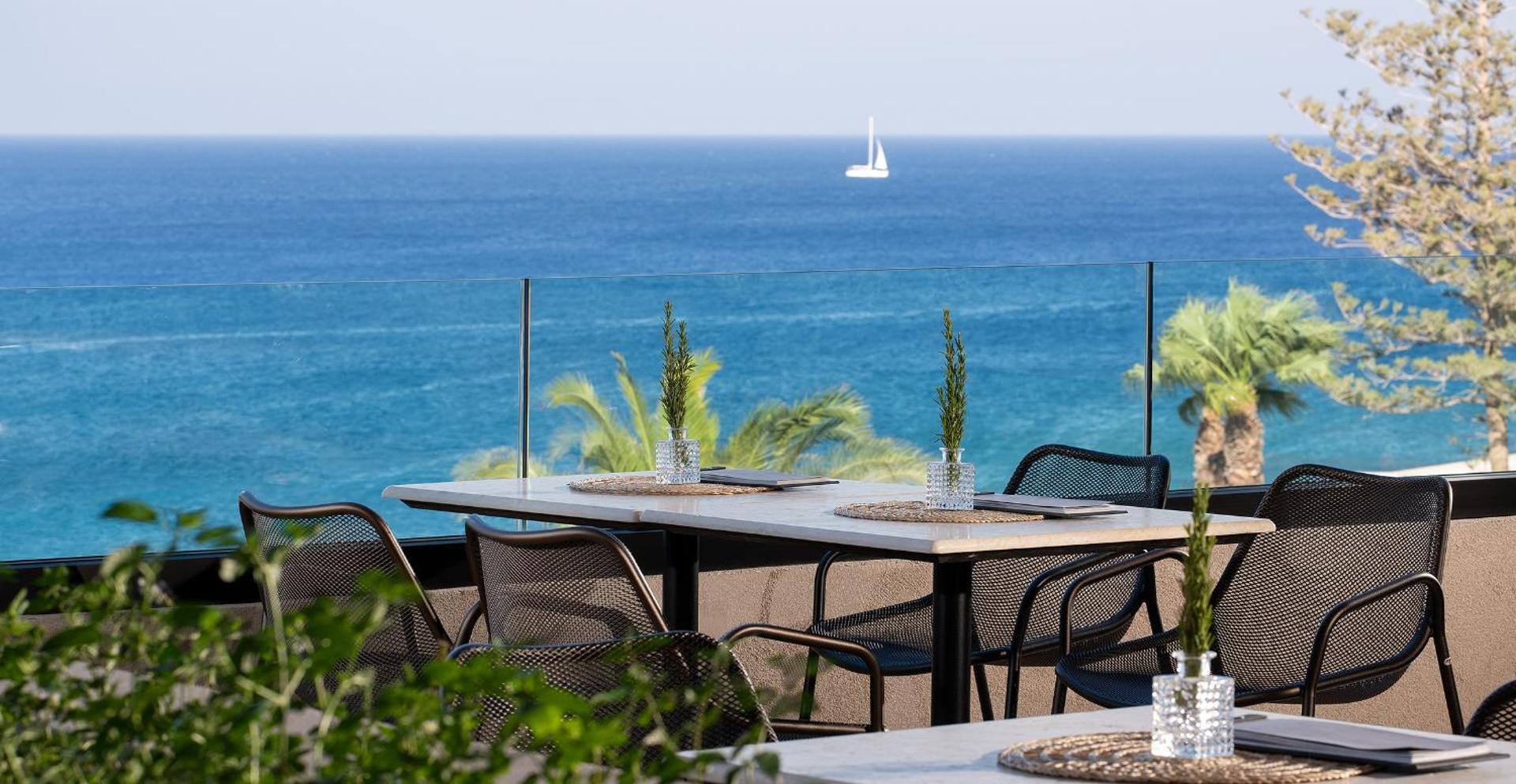 Helea Lifestyle Beach Resort restorāna terase Rodas salā ar skatu uz jūru, galdiem un melniem krēsliem pie stikla margām