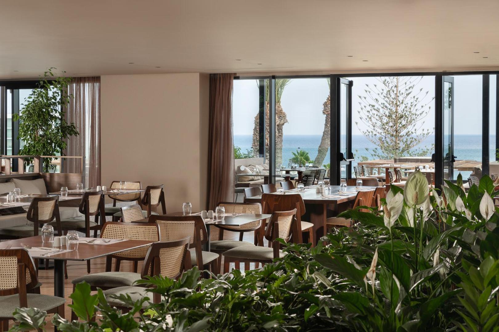 Helea Lifestyle Beach Resort moderns restorāns ar koka mēbelēm, telpaugiem un lieliem logiem, kas paver skatu uz palmu dārzu