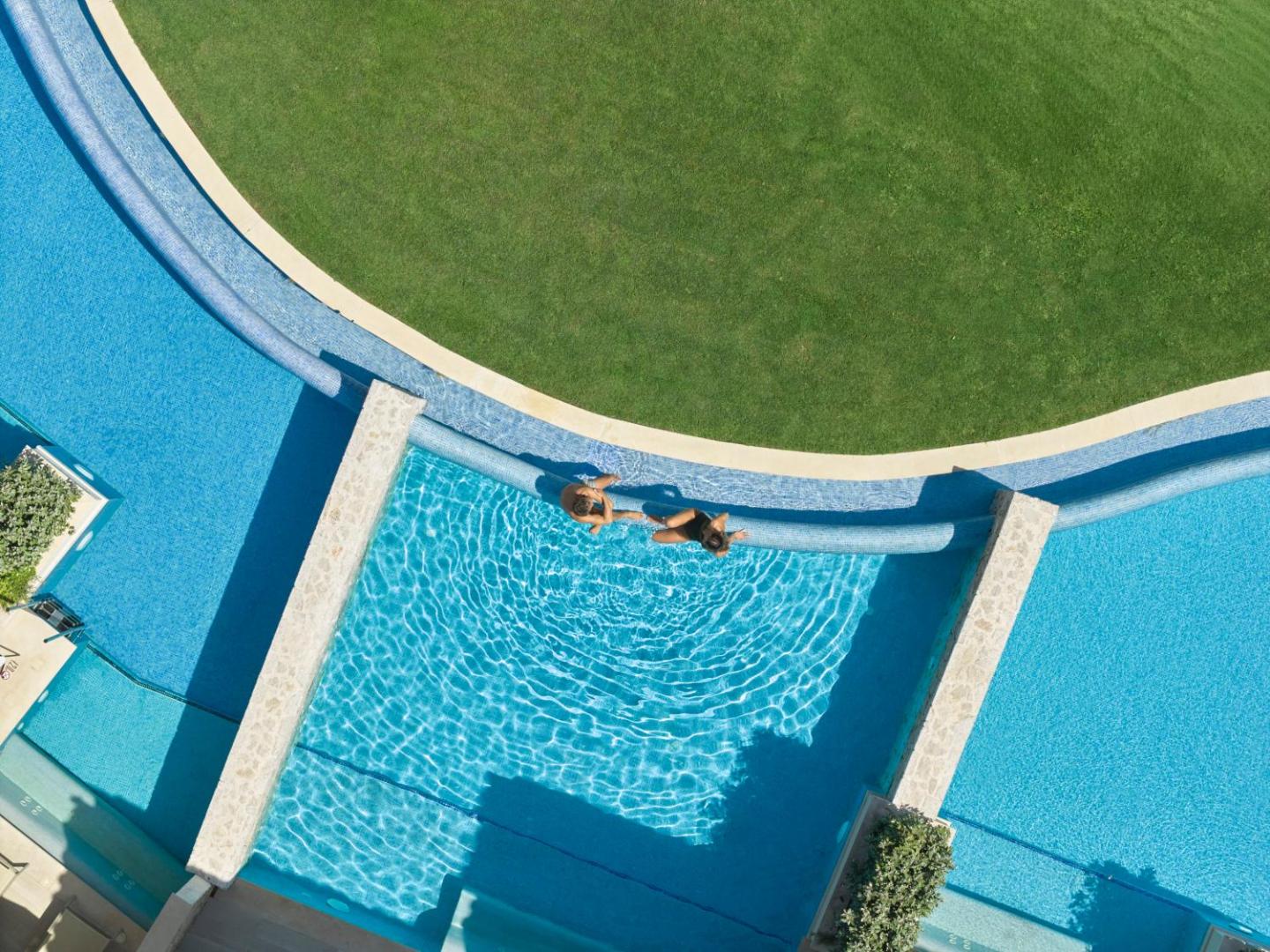 Skats no augšas uz Atlantica Imperial Resort baseinu ar pārplūdes malu, kur divi cilvēki atpūšas pie ūdens un zaļojoša zālāja.