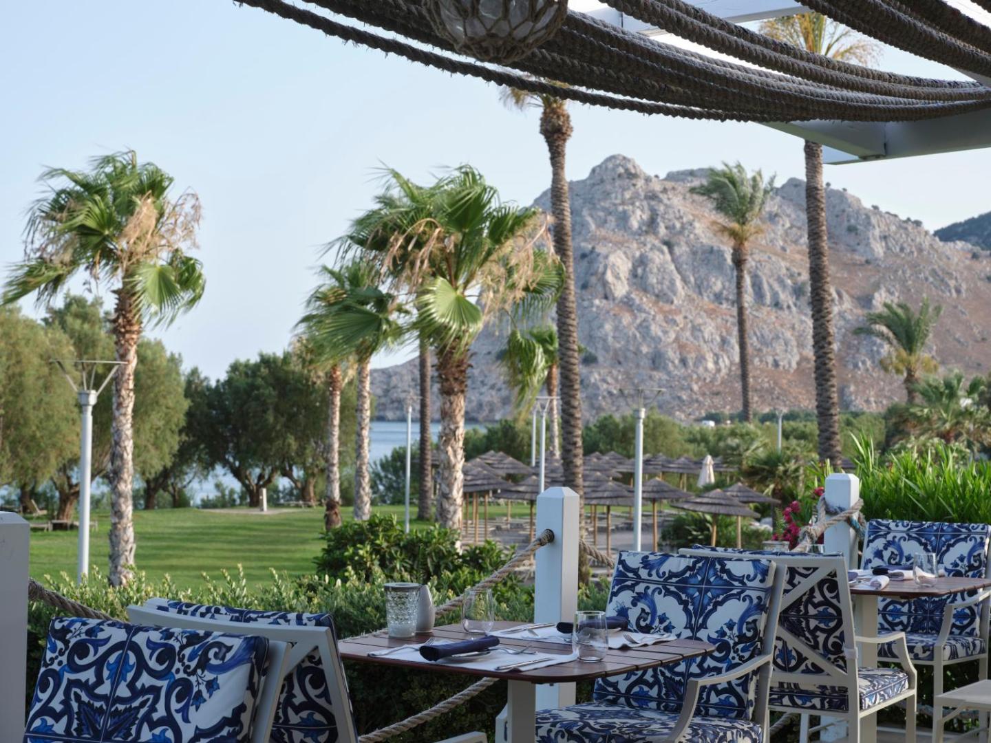 Āra terase Atlantica Imperial Resort ar skatu uz palmām un kalnu, galdi klāti ar ziliem rakstainiem krēsliem