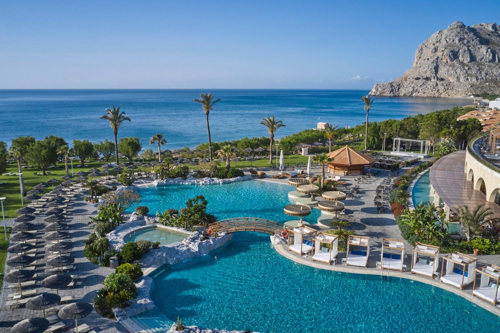 Atlantica Imperial Resort Rodā panorāmas skats uz plašo baseinu kompleksu, Vidusjūras piekrasti un apkārtējiem kalniem saulainā dienā.