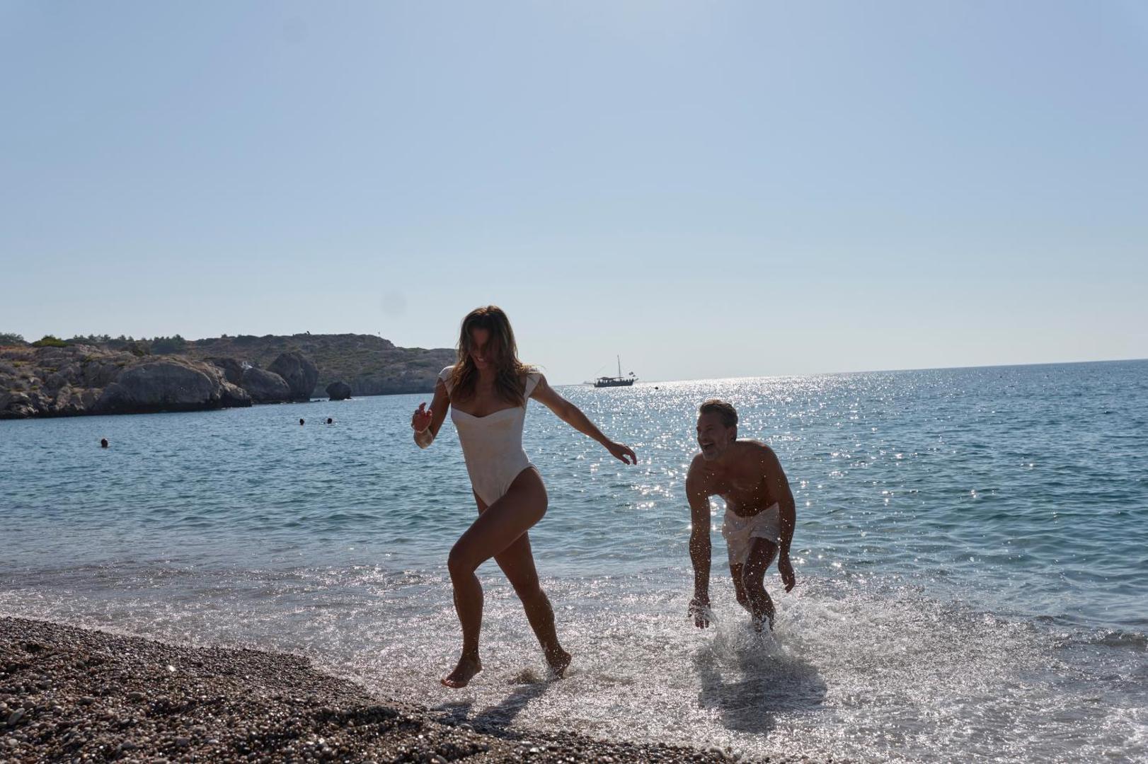 Pāris skrien pa Atlantica Imperial Resort pludmali Rodas salā — saulaina diena pie jūras, ūdens šļakatas un kalnains fons