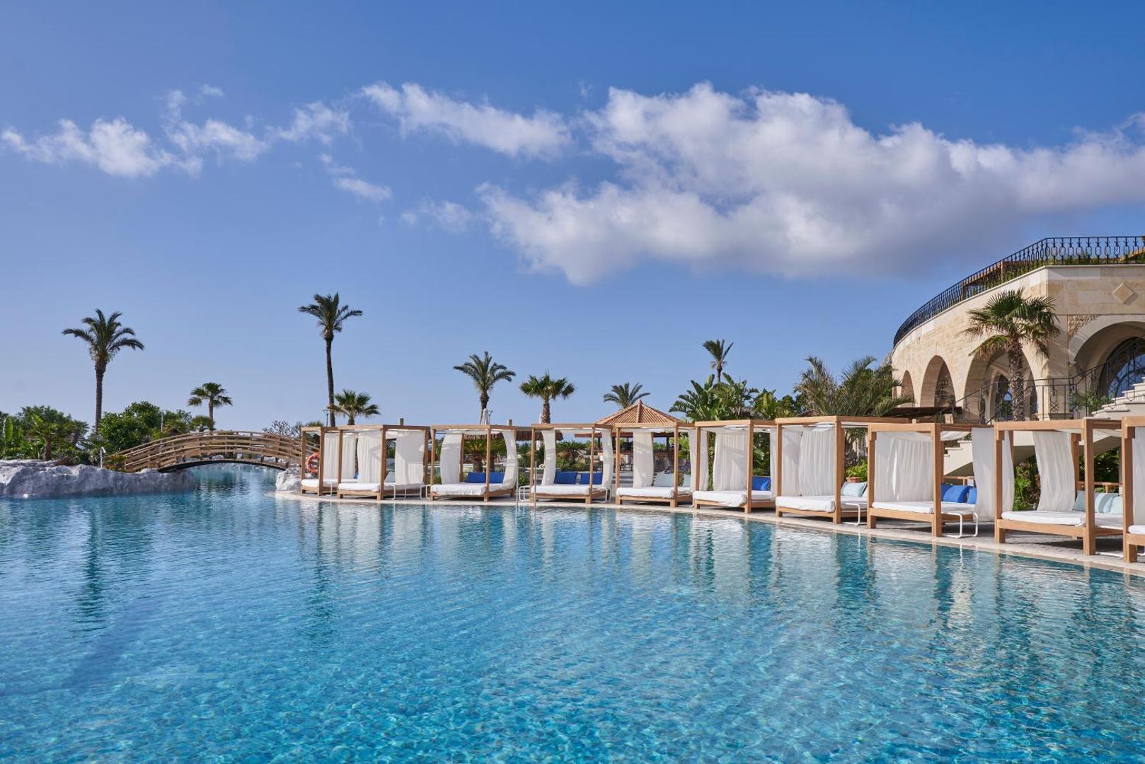 Atlantica Imperial Resort baseina mala ar greznām atpūtas gultām, koka tiltiņu un viesnīcas galveno ēku fonā zem skaidrām debesīm.