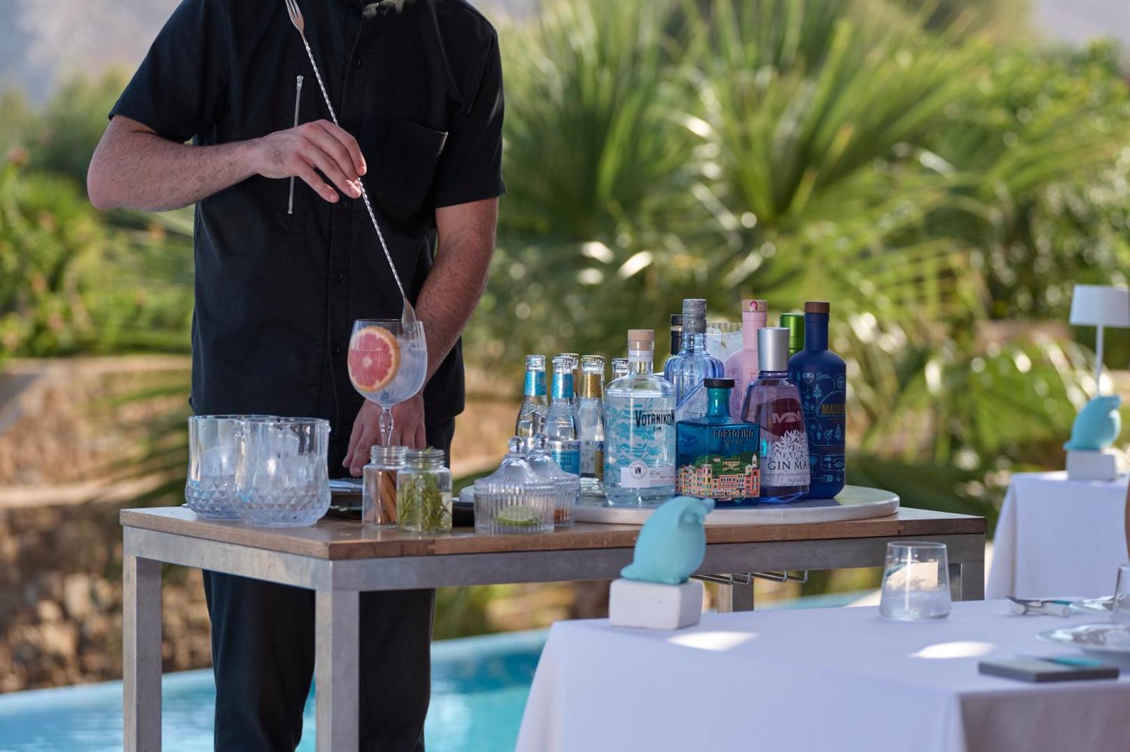 Bārmenis gatavo kokteili Atlantica Imperial Resort pie baseina, fonā redzamas dažādas džina pudeles un tropiski apstādījumi Rodā