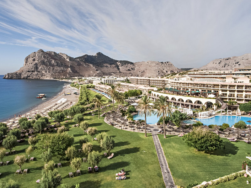 Atlantica Imperial Resort zaļā dārza zona ar palmām, sauļošanās krēsliem zālienā un skatu uz pludmali un kalnu grēdu
