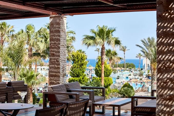 Atlantica Aegean Blue viesnīcas āra terase ar skatu uz baseinu un jūru Rodas salā — atpūtas zona ar mīkstajiem krēsliem un palmām