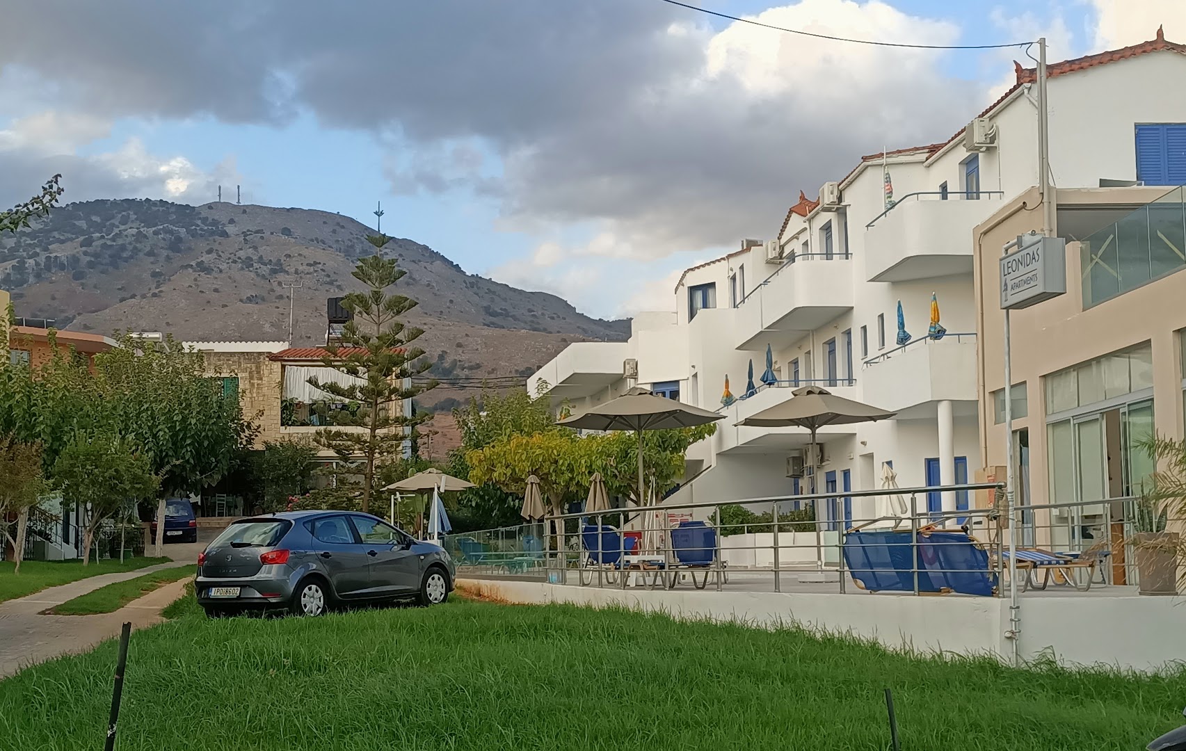Leonidas Apartments ēka Mare Village kompleksā ar balkoniem un saulessargiem — skats uz kalniem un zaļo zonu pie viesnīcas