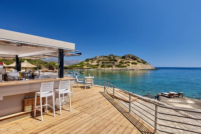 Blue Marine Resort & Spa āra bārs uz koka terases ar skatu uz jūru un kalnainu piekrasti Krētas salā