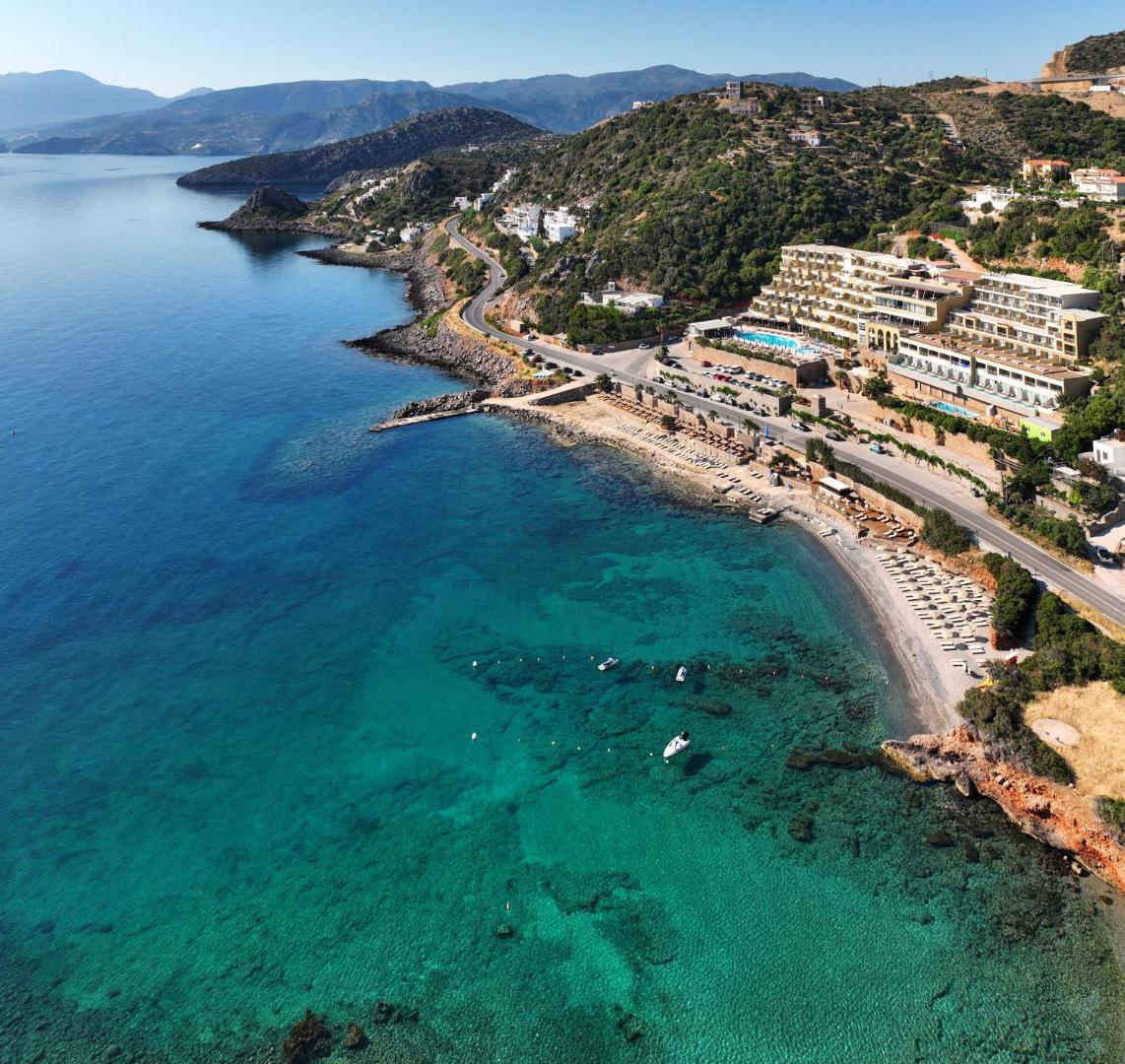 Blue Marine Resort & Spa panorāmas skats no putna lidojuma uz viesnīcas kompleksu, privāto pludmali un tirkīzzilo Mirabello līča ūdeni Krētā