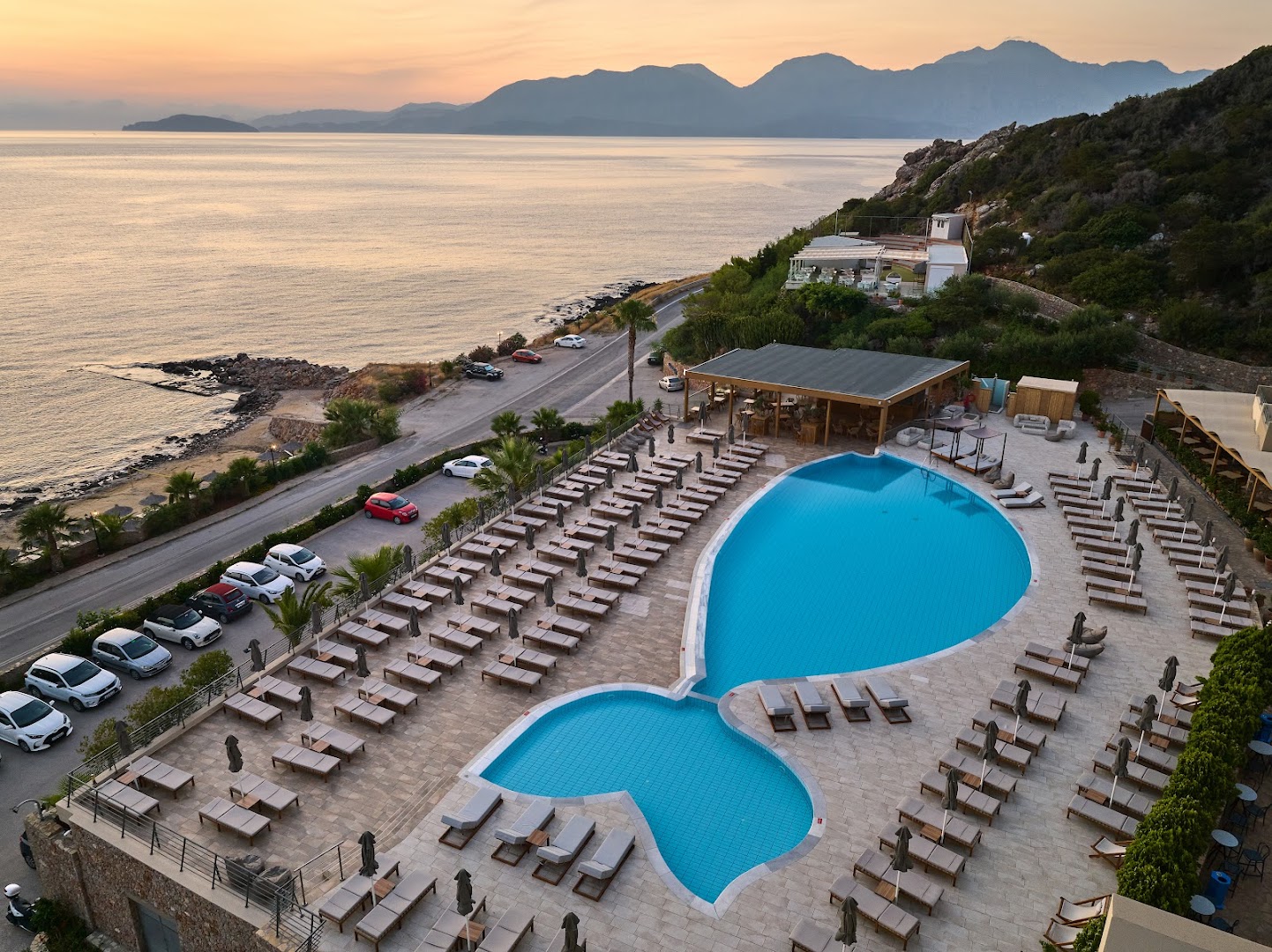 Blue Marine Resort & Spa zivs formas baseins no augšas saulrieta laikā, blakus esošā piekrastes josla un kalnu silueti Krētā