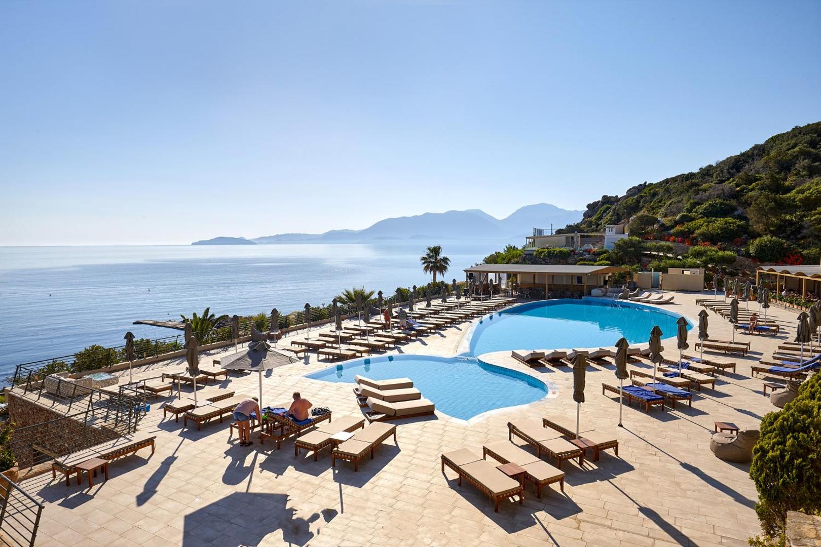 Blue Marine Resort & Spa āra baseins ar skatu uz Mirabello līci Krētā, saulainā dienā ar atpūtas krēsliem un kalniem fonā