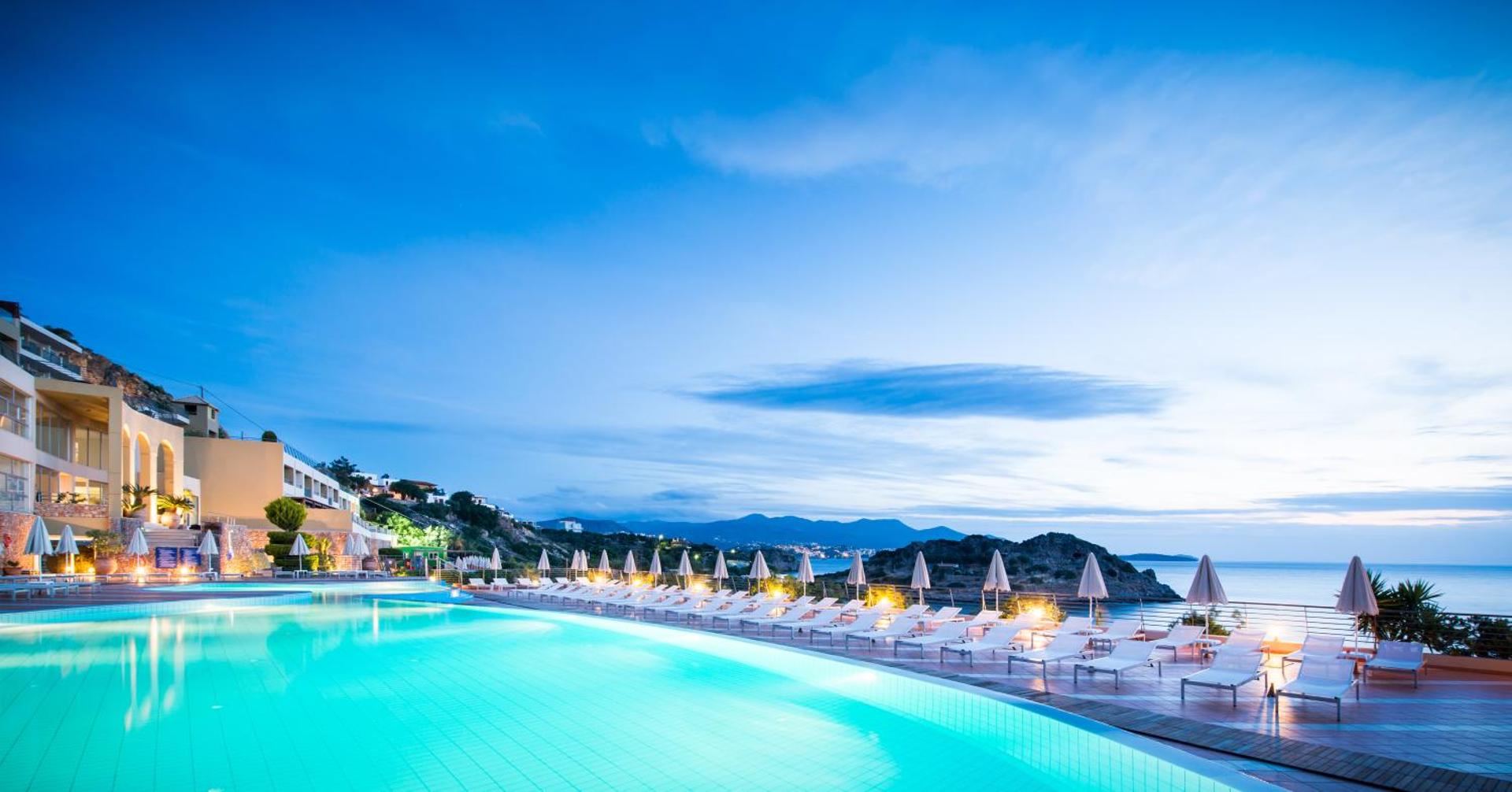 Blue Marine Resort & Spa izgaismots baseins krēslas stundā ar skatu uz piekrasti un kalniem, moderni sauļošanās krēsli un viesnīcas korpuss