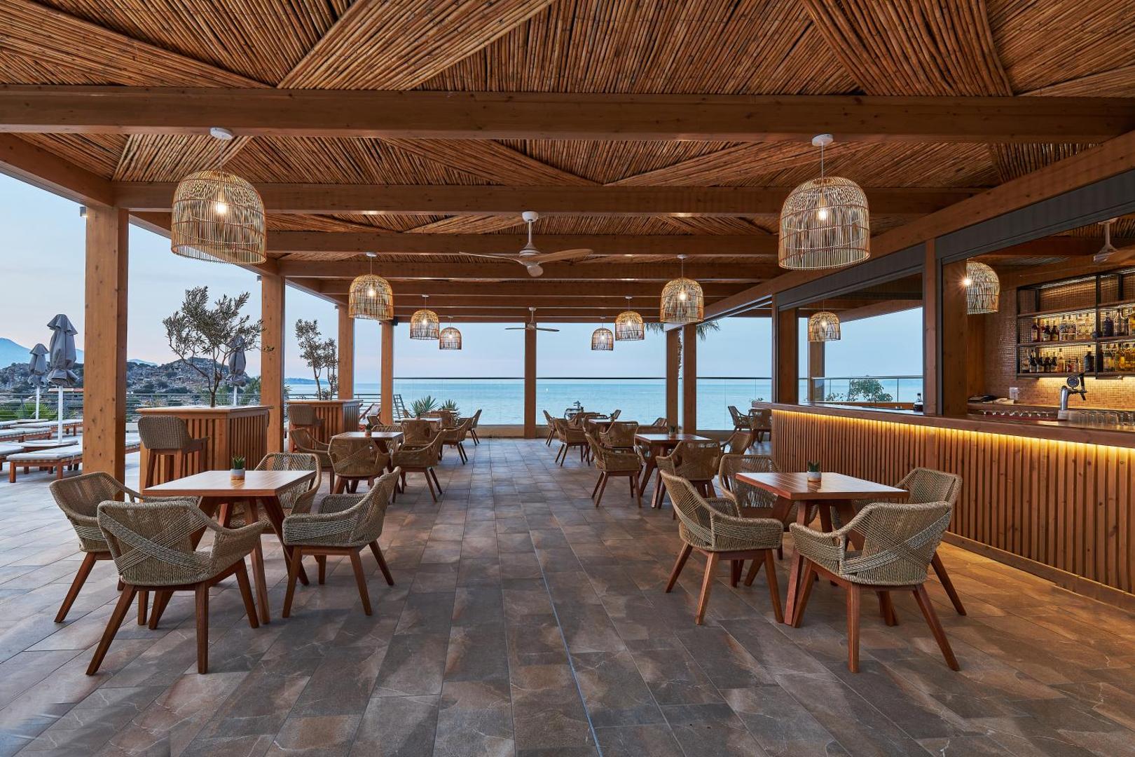 Blue Marine Resort & Spa atvērta tipa terases bārs ar pītiem krēsliem un skatu uz jūru saulrietā Krētas piekrastē.