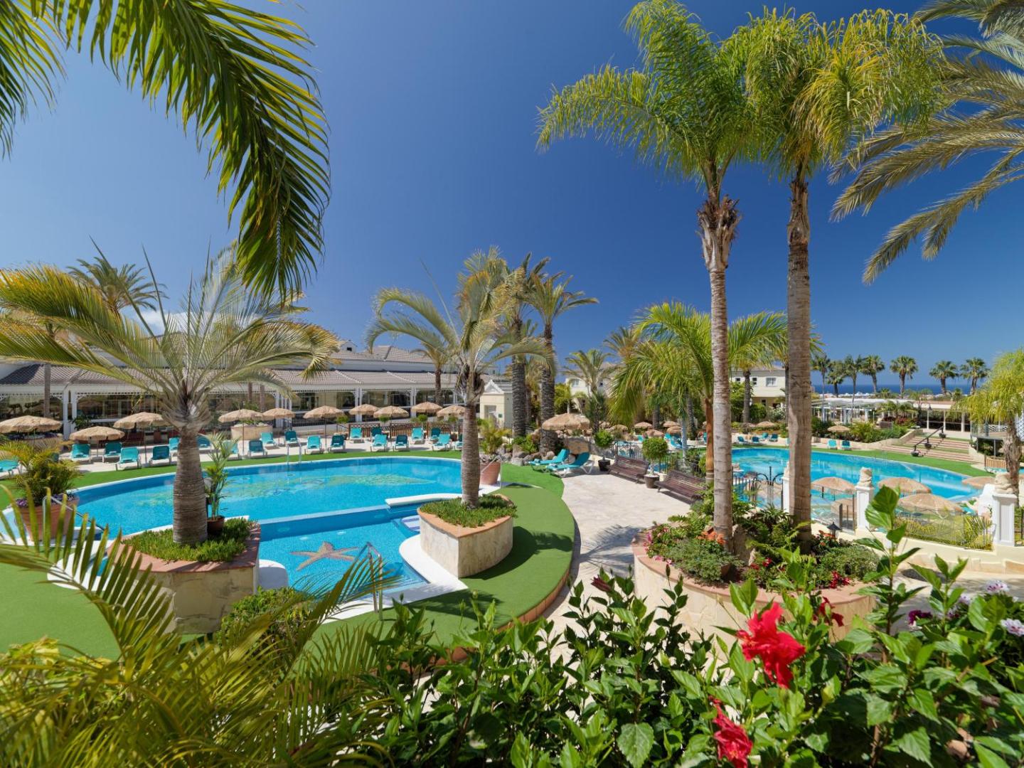 Gran Oasis Resort baseinu komplekss ar palmām un ziedošiem krūmiem Tenerifē — skats caur tropu augiem uz atpūtas zonu