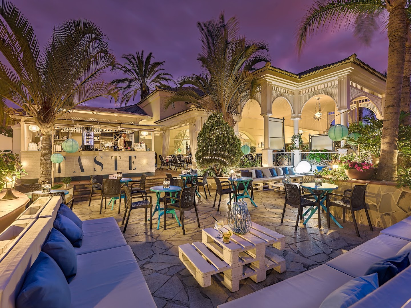 Gran Oasis Resort āra terase Taste ar bāru, galdiņiem un palmām vakara apgaismojumā Tenerifes dienvidos piekrastes tuvumā