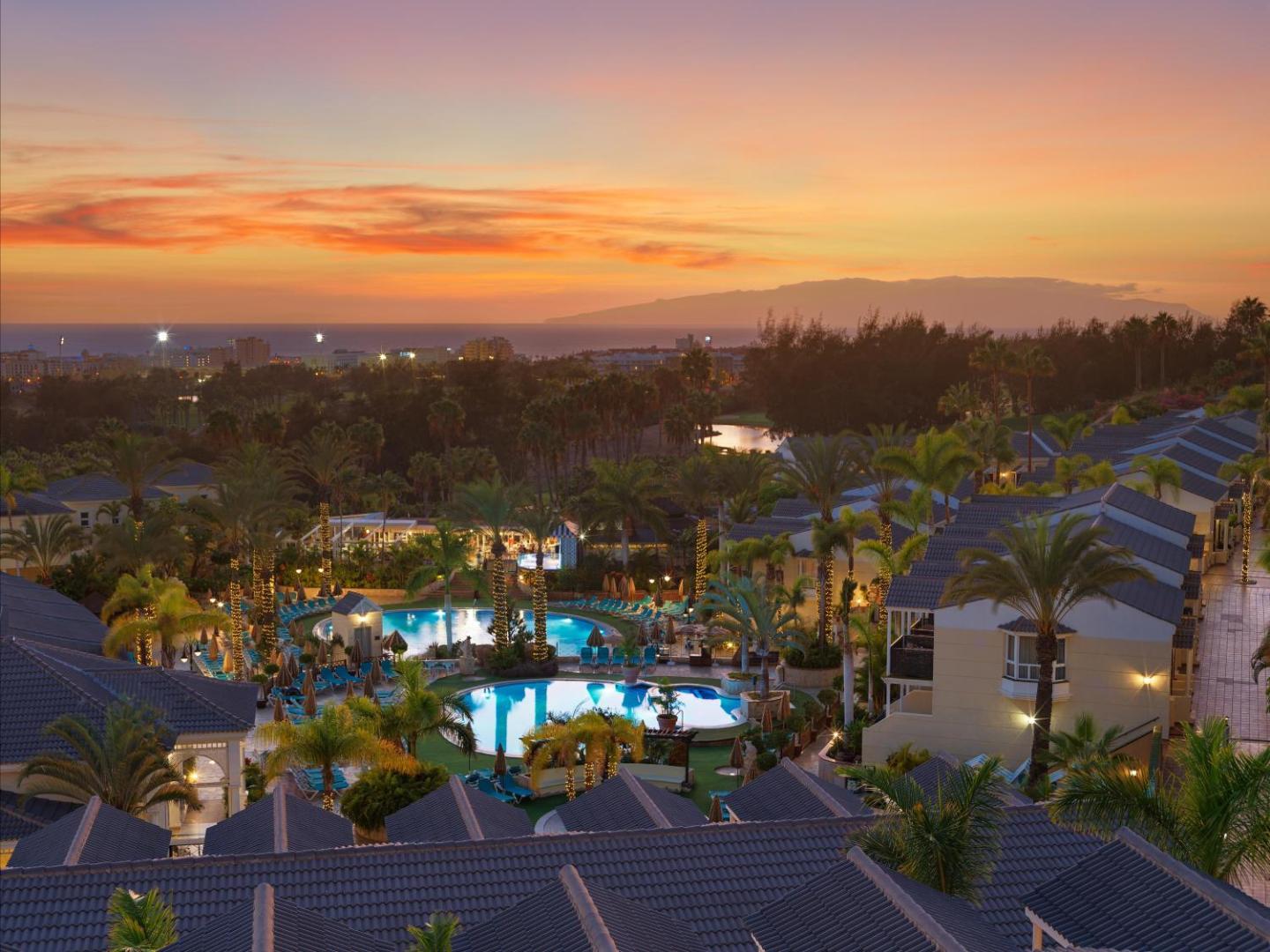 Gran Oasis Resort panorāmas skats saulrietā pār viesnīcas teritoriju un okeānu Tenerifē — baseini, bungalo tipa ēkas un kalni tālumā