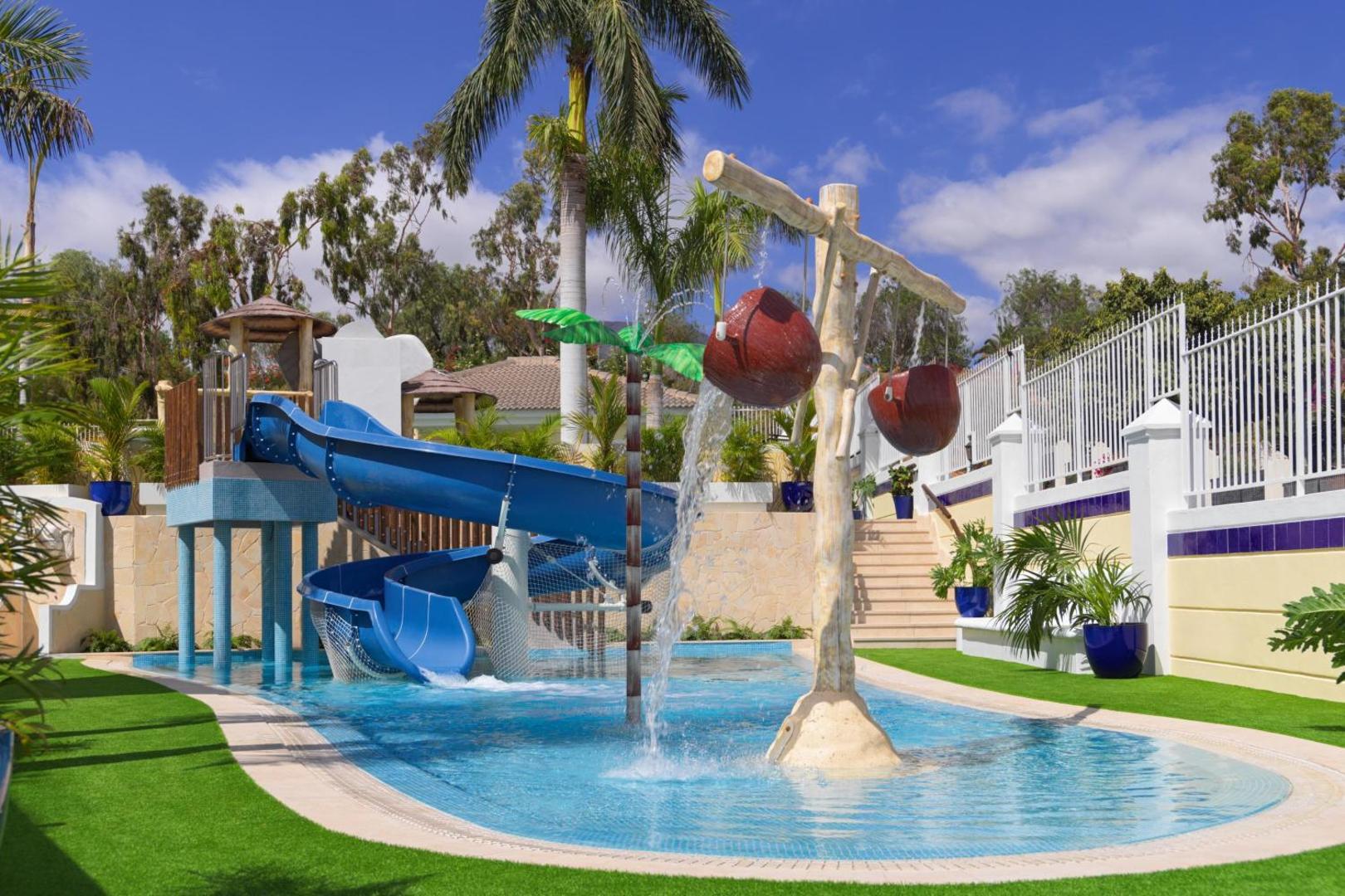 Gran Oasis Resort bērnu baseins ar zilu ūdens slidkalniņu un dekoratīvām ūdens strūklām Tenerifē — apkārt zaļš zāliens un palmas