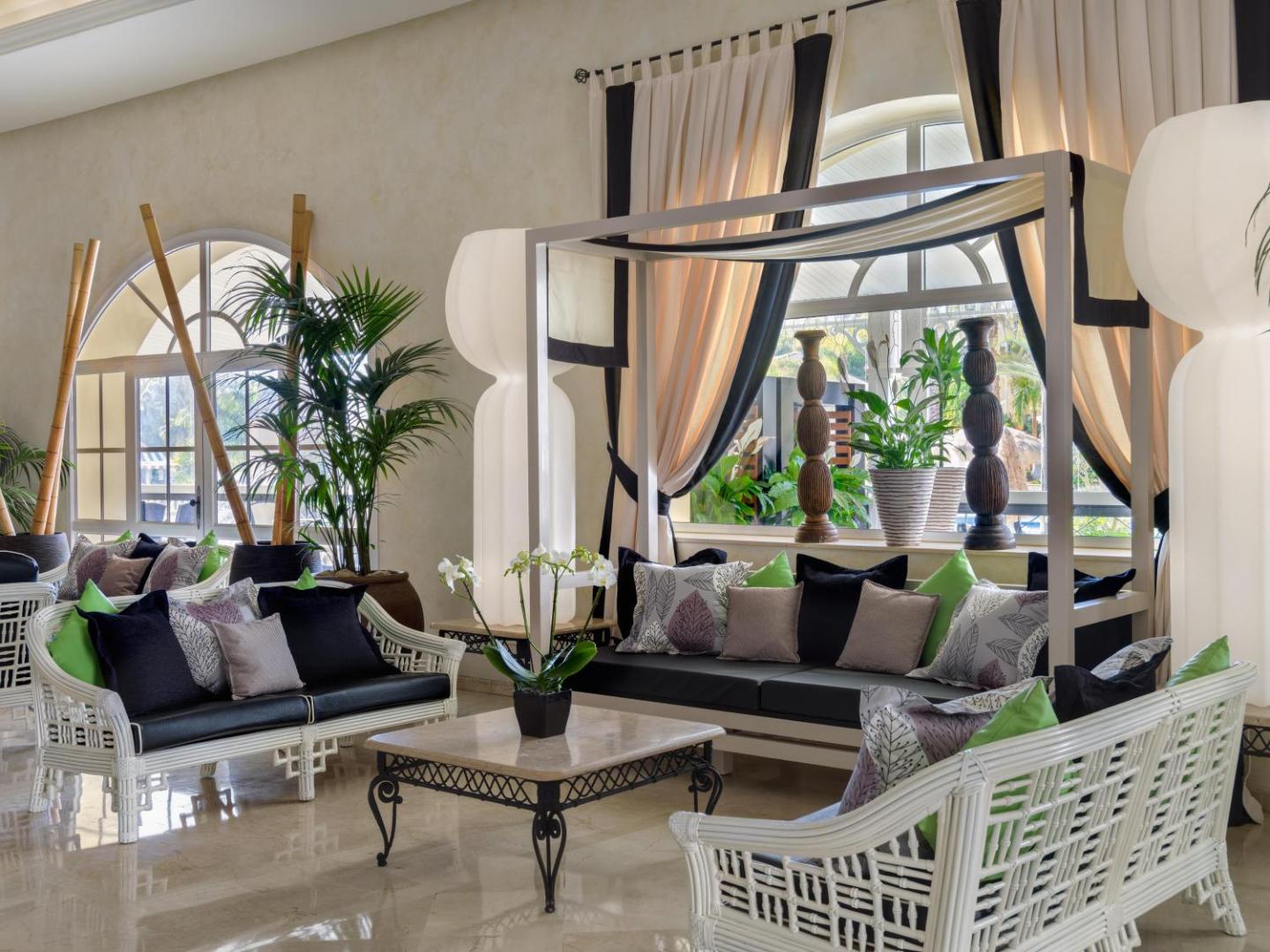 Gran Oasis Resort viesnīcas lobby atpūtas zona Tenerifē ar gaišiem pītiem dīvāniem, dekoratīviem spilveniem un eksotiskiem telpaugiem mājīgā atmosfērā