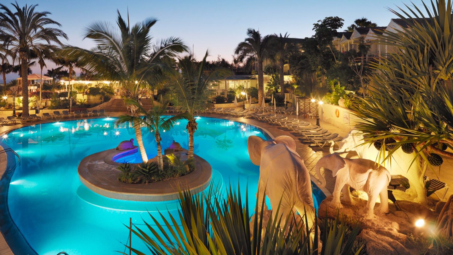 Gran Oasis Resort izgaismots āra baseins vakarā ar ziloņu statujām un palmām Tenerifē, Kanāriju salās — romantiska atmosfēra un atpūtas zona