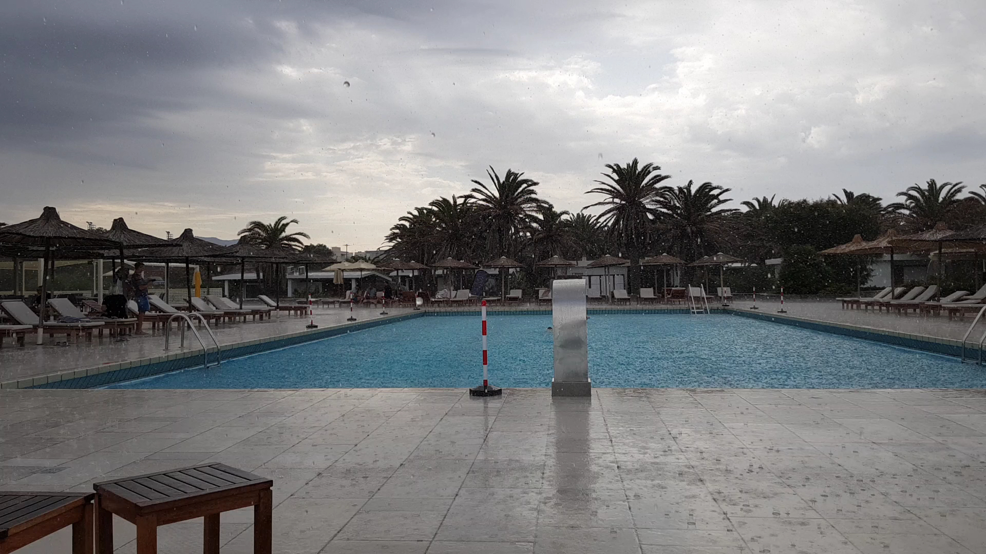 Civitel Creta Beach plašais āra baseins lietus laikā ar palmām fonā un tukšiem sauļošanās krēsliem Krētas kūrortā