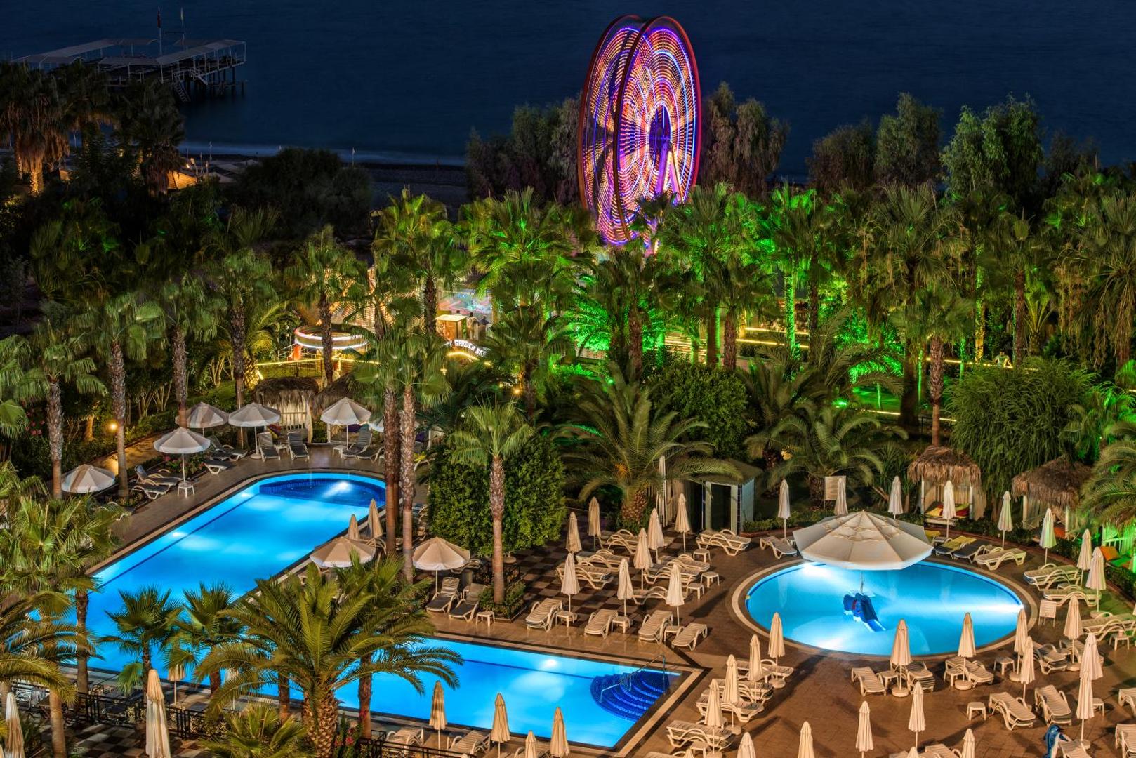 Delphin Deluxe viesnīcas vakara skats uz izgaismotiem baseiniem, palmām un panorāmas ratu Alānijas reģionā, Turcijā
