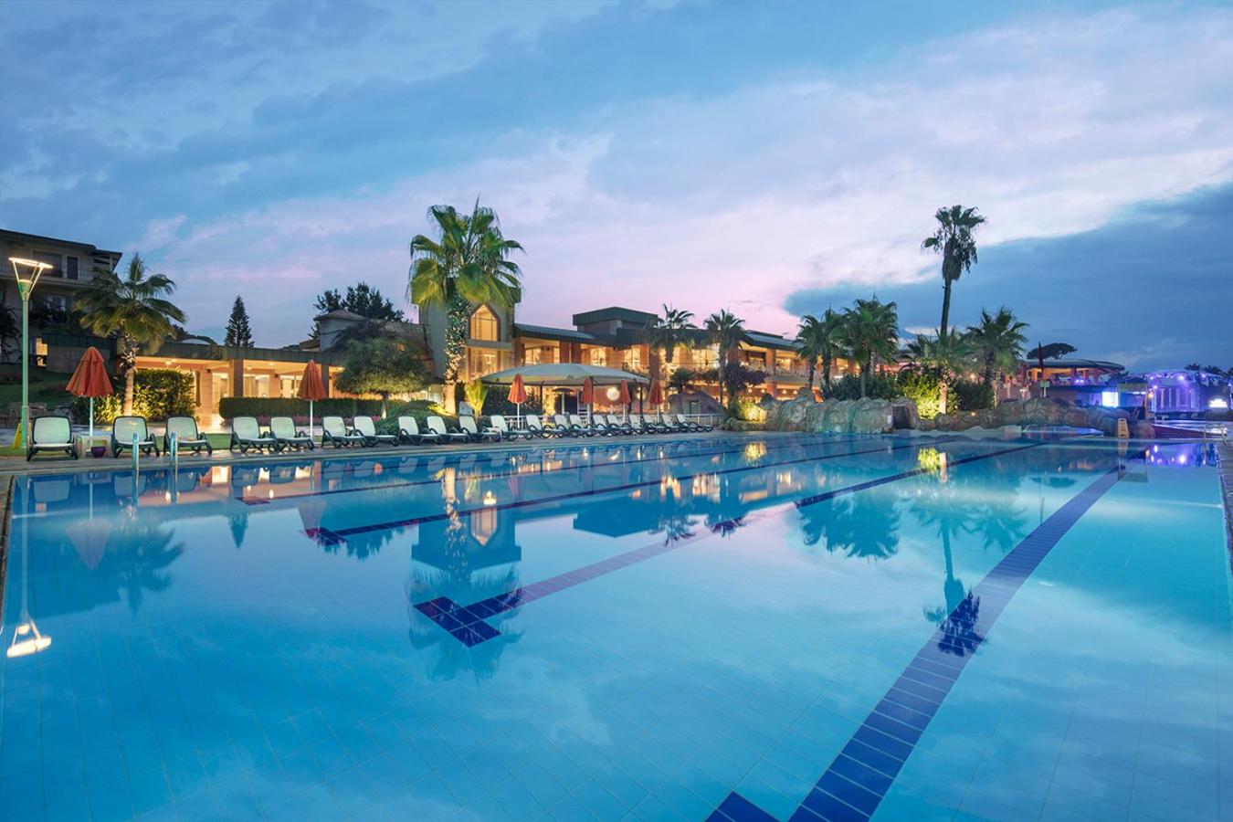 Pine Beach Belek plašais āra baseins saulrieta laikā ar palmām, atpūtas krēsliem un viesnīcas korpusu fonā