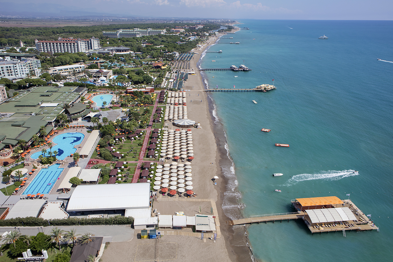 Panorāmas skats uz Pine Beach Belek kūrorta piekrasti, rādot baseinu kompleksu, pludmales zonu un Vidusjūras dzidro ūdeni.